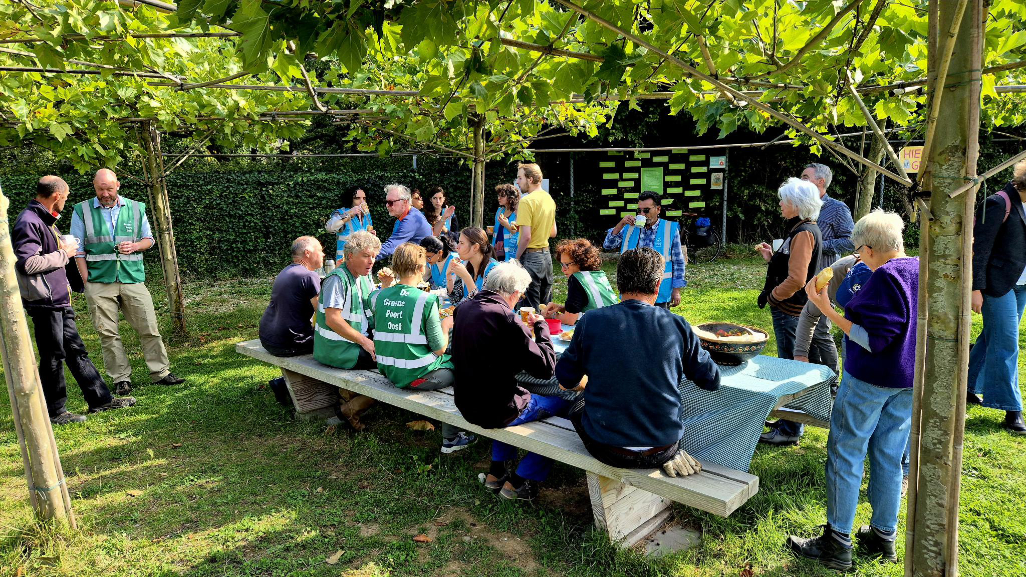20 vrijwilligers zitten en staan rondom de grote lunchtafel op het gras tussen de platanen.